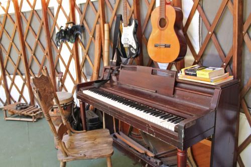 A wooden piano with a chair beside it, surrounded by guitars and musical instruments in a cozy, rustic setting.
