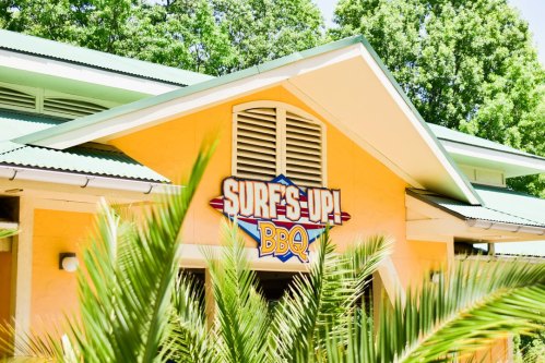 A colorful building with a sign that reads "Surf's Up! BBQ," surrounded by lush green foliage.