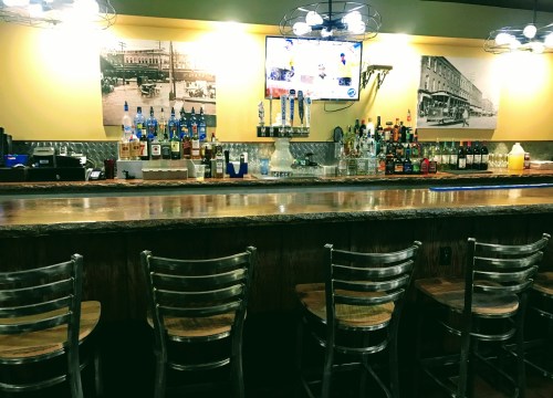 A bar with a wooden counter, metal stools, and shelves stocked with various bottles, plus a TV on the wall.
