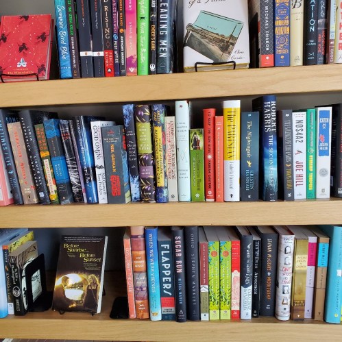 A wooden bookshelf filled with various colorful books, showcasing a mix of genres and authors.