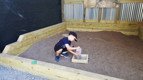 A young boy in a cap plays in a sandy area, using a container to sift through the sand.