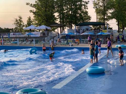 A lively water park scene with children playing in a wave pool, surrounded by trees and lounge chairs.