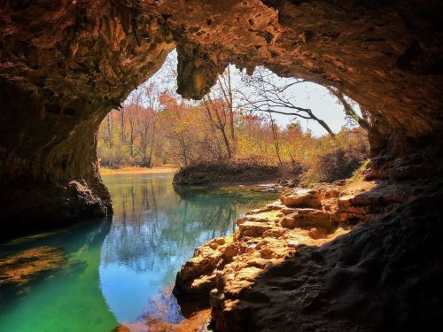 A serene cave opening reveals a tranquil river surrounded by autumn foliage and rocky terrain.
