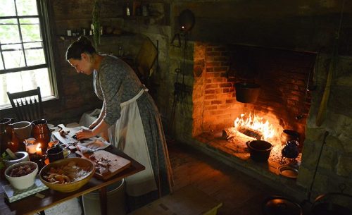 A woman in period clothing prepares food at a rustic kitchen table near a roaring fireplace.