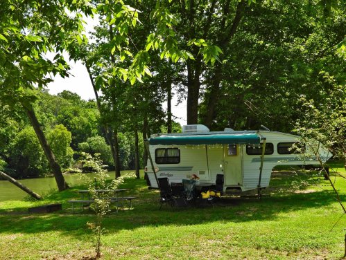 A vintage camper parked by a river, surrounded by lush green trees and a picnic area with chairs.