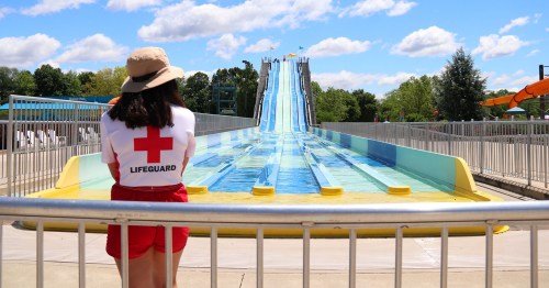 A lifeguard in a red and white uniform watches over a water slide at a sunny water park.