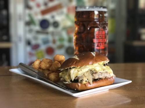 A pretzel bun sandwich with pork and sauerkraut, served with tater tots and a large beer in the background.