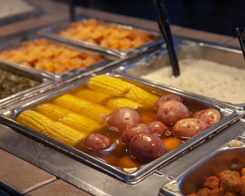 A buffet display featuring corn on the cob and red potatoes in a metal tray, with other food items in the background.