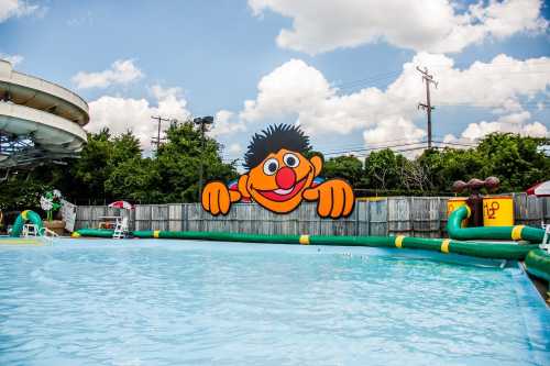 A colorful cartoon character peeks over a swimming pool with water slides in the background under a blue sky.