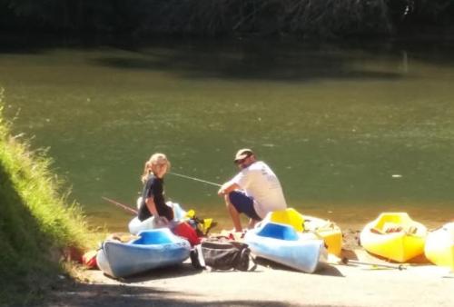 Two people sit by a river, preparing kayaks and fishing gear on the shore. Bright kayaks are lined up nearby.