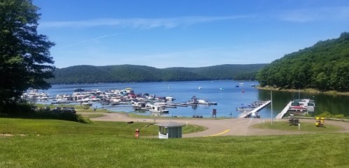 A scenic view of a marina on a calm lake, surrounded by green hills under a clear blue sky.