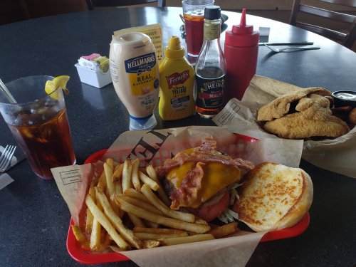 A burger with bacon and cheese, fries, and condiments on a table with a drink and fried food in the background.