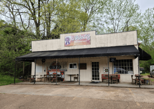 A small, rustic restaurant with a sign reading "Ubon's" and outdoor seating under a black awning, surrounded by trees.