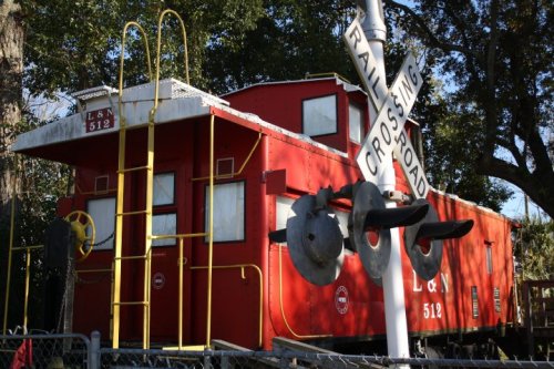 A red caboose labeled "L&N 512" next to a railroad crossing sign, surrounded by trees.
