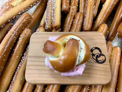 A soft pretzel sandwich with pink filling on a wooden board, surrounded by long pretzel sticks.