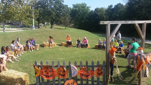 A group of children and adults sit on hay bales in a circle, surrounded by Halloween decorations in a park.