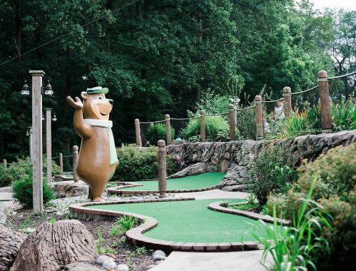 A cartoon bear statue waves beside a mini-golf course, surrounded by greenery and stone pathways.