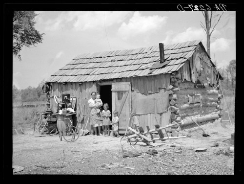 A family stands outside a small, rustic wooden cabin with a tin roof, surrounded by a rural landscape.