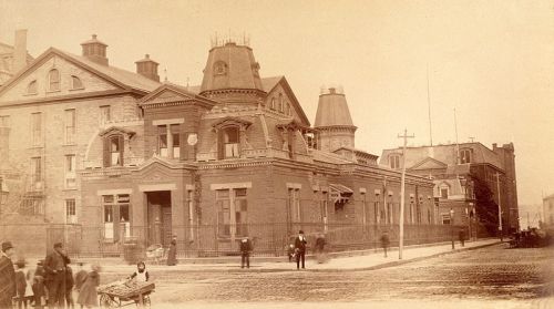 Historic sepia-toned photo of a grand building with ornate architecture, surrounded by people and cobblestone streets.
