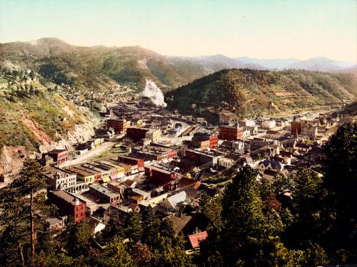 Aerial view of a colorful historic town nestled in mountains, with buildings, streets, and greenery visible.