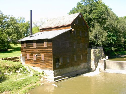 A historic wooden mill building beside a river, featuring a stone foundation and a chimney, surrounded by greenery.