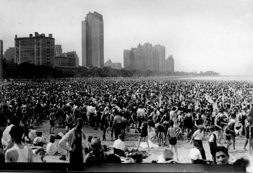 A crowded beach scene with numerous people swimming and sunbathing, surrounded by tall buildings in the background.