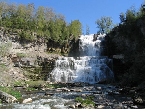 A cascading waterfall flows over rocky cliffs, surrounded by lush greenery and a clear blue sky.