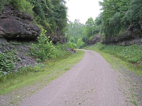 A gravel path winds between rocky cliffs, surrounded by lush greenery and trees under a cloudy sky.