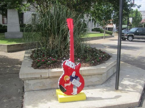 A large, red guitar sculpture with colorful designs, placed in a landscaped area near a street.