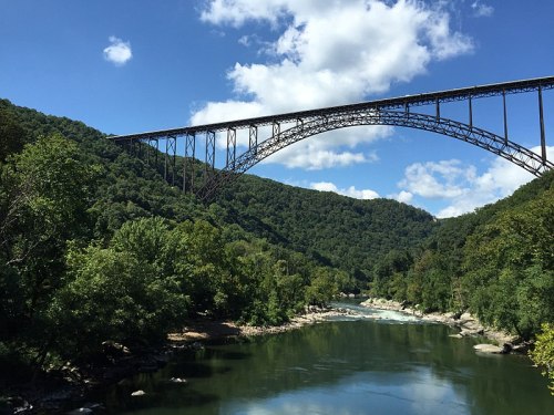 A tall metal bridge arches over a river, surrounded by lush green hills and a blue sky with fluffy clouds.
