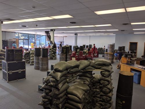A spacious room filled with stacked sandbags and barrels, with people organizing equipment in the background.