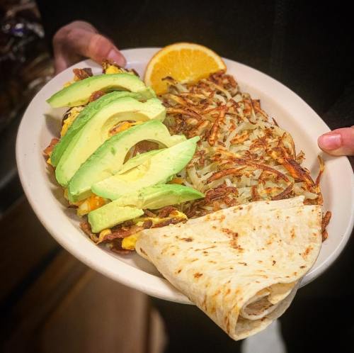 A plate of breakfast featuring avocado slices, hash browns, a tortilla, and a slice of orange.