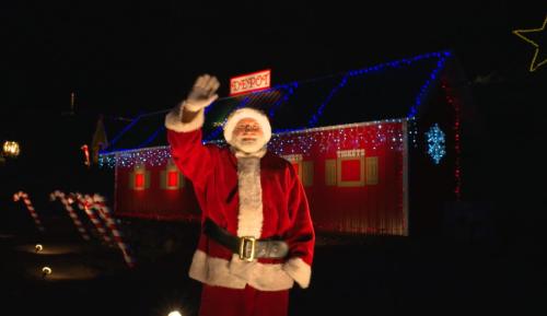 A cheerful Santa Claus waves in front of a brightly lit, festive building decorated for the holidays.
