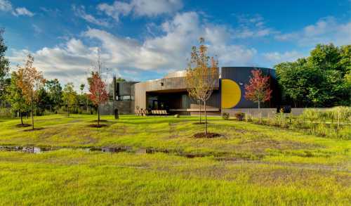 Modern building with a circular design, surrounded by green grass and trees under a blue sky with scattered clouds.