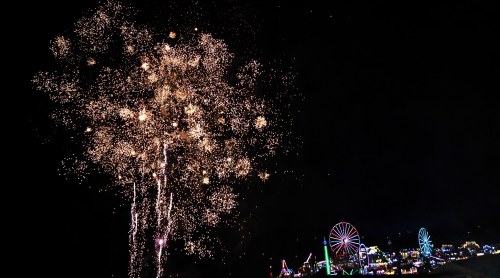 Colorful fireworks illuminate the night sky above a vibrant carnival with lights and a Ferris wheel.