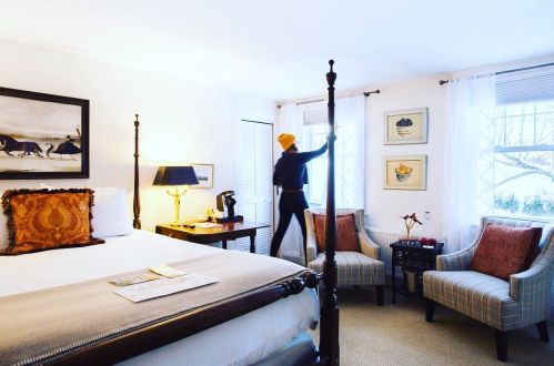 A cozy hotel room with a bed, armchairs, and a person adjusting the curtains by the window.