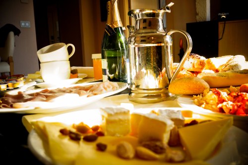 A table spread with various cheeses, meats, pastries, a silver pitcher, and bottles of champagne.