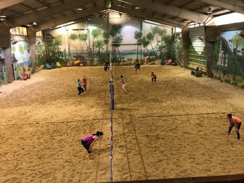 Indoor sand volleyball court with players in action, surrounded by tropical-themed murals.