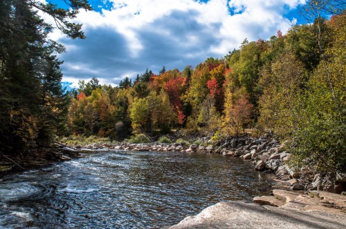 A serene river flows through a forest with vibrant autumn foliage under a partly cloudy sky.