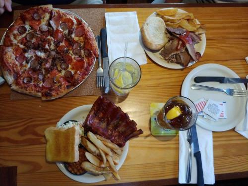 A table spread with a large pizza, a plate of ribs, fries, toast, and drinks, including a glass of lemonade and iced tea.
