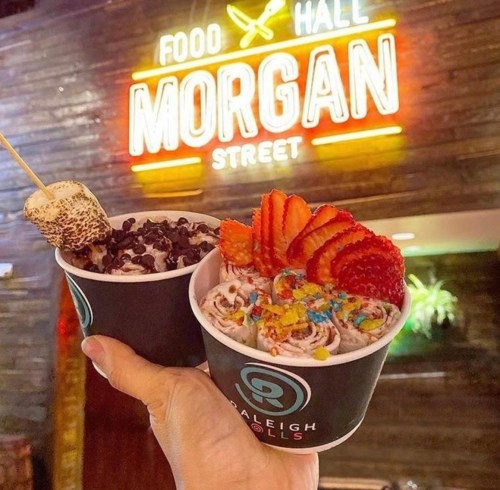 A hand holds two cups of rolled ice cream topped with strawberries, sprinkles, and a marshmallow, with a neon sign in the background.