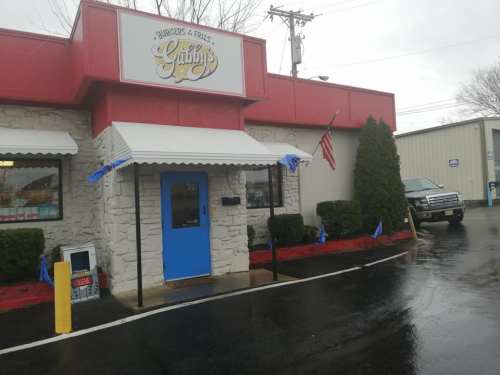 Exterior of Gabby's Burgers & Fries, featuring a blue door, flags, and a parked truck on a rainy day.