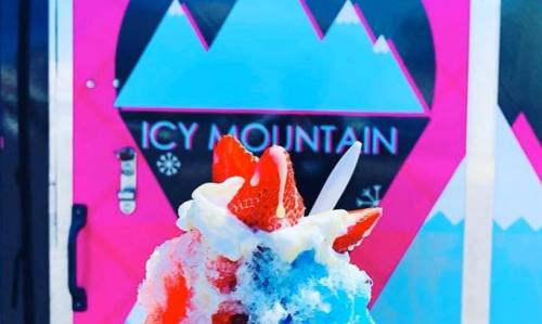 A colorful snow cone topped with strawberries in front of a vibrant "Icy Mountain" shop sign.