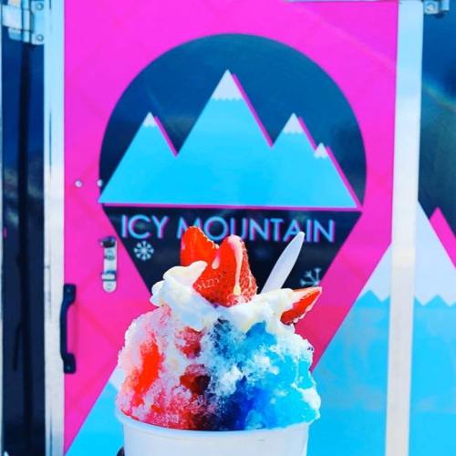 A colorful snow cone topped with whipped cream and strawberries, in front of a pink and black "Icy Mountain" stand.