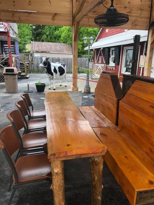 A rustic wooden table with chairs under a shelter, with a cow statue in the background near a red building.