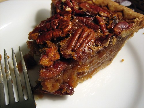 A slice of pecan pie on a white plate, with a fork resting beside it. The pie has a glossy, nutty topping.