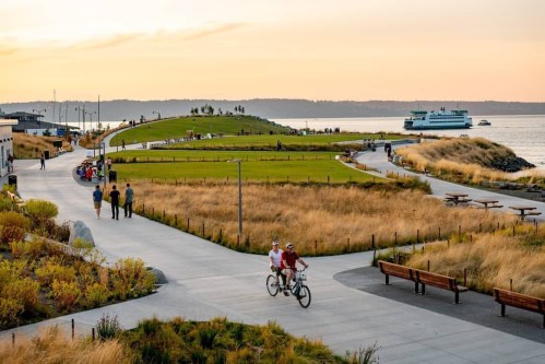 A scenic waterfront park with people walking and biking, surrounded by grass and benches, under a sunset sky.