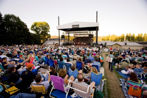 A large outdoor concert with a crowd seated on blankets and chairs, enjoying a performance on stage.