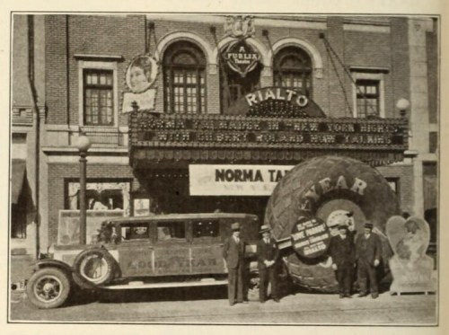 Historic photo of the Rialto Theatre with a large tire display and people in vintage clothing outside.