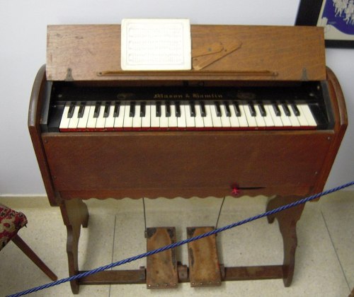 A vintage wooden harmonium with a keyboard, foot pedals, and sheet music holder, displayed in a room.